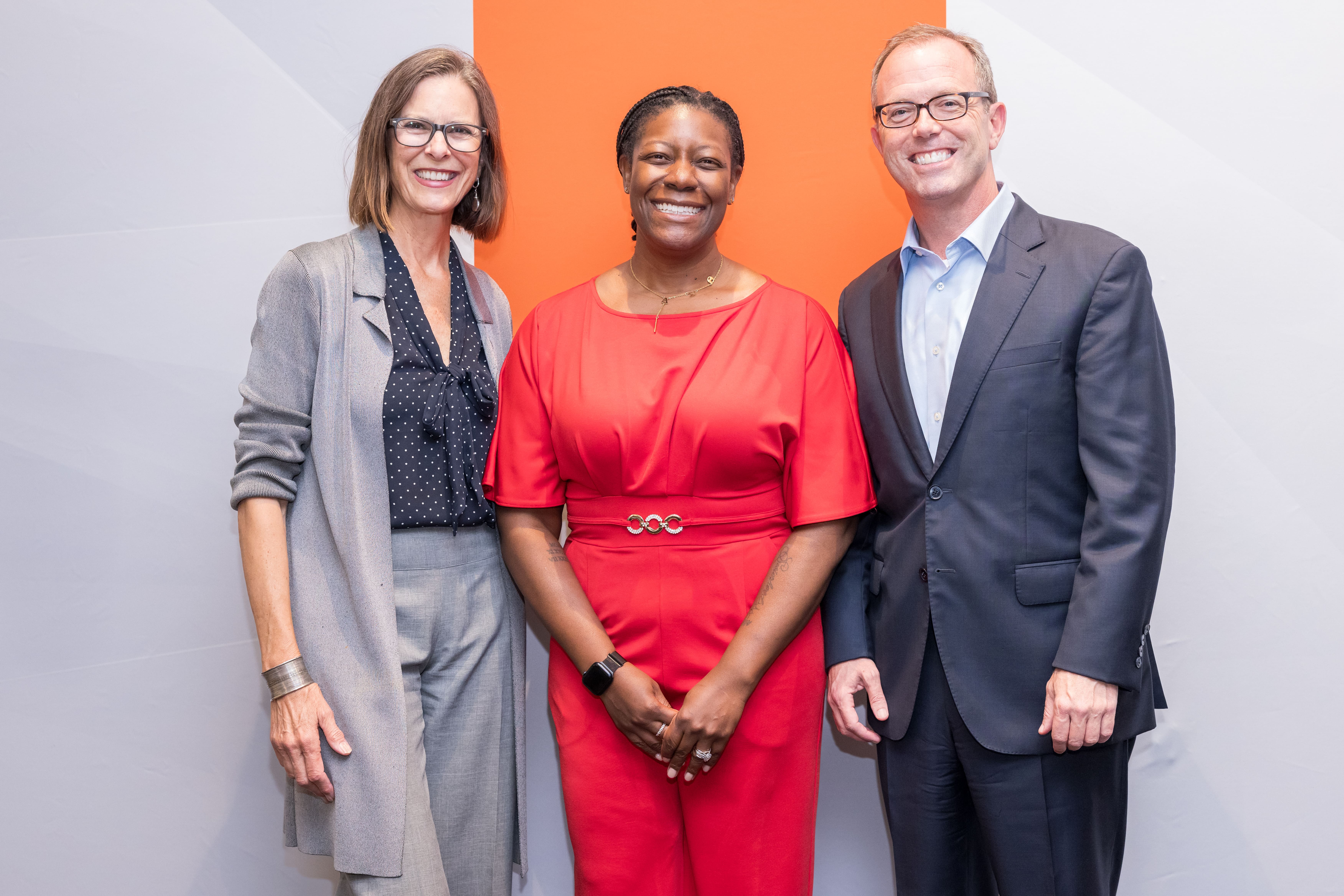 Three people smiling and posing together against a white and orange background at an event.
