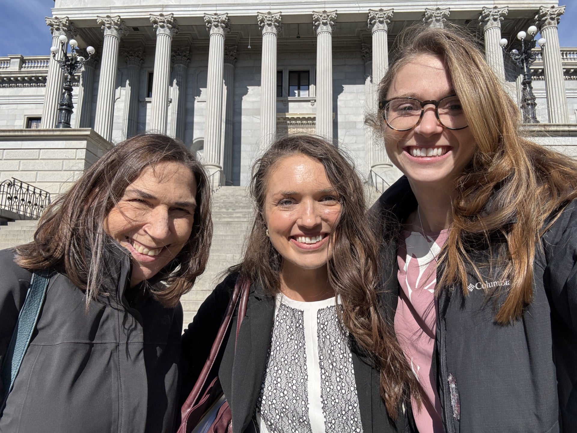 Three people smiling for a selfie in front of a large building with tall columns and steps.