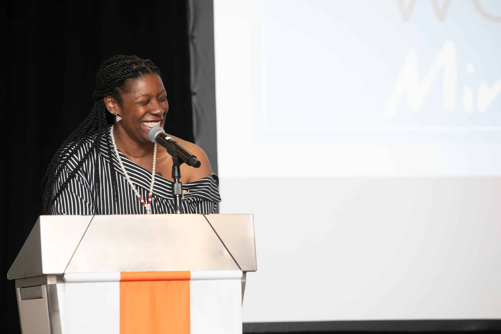 Speaker smiling while speaking into a microphone at a podium during an event presentation.