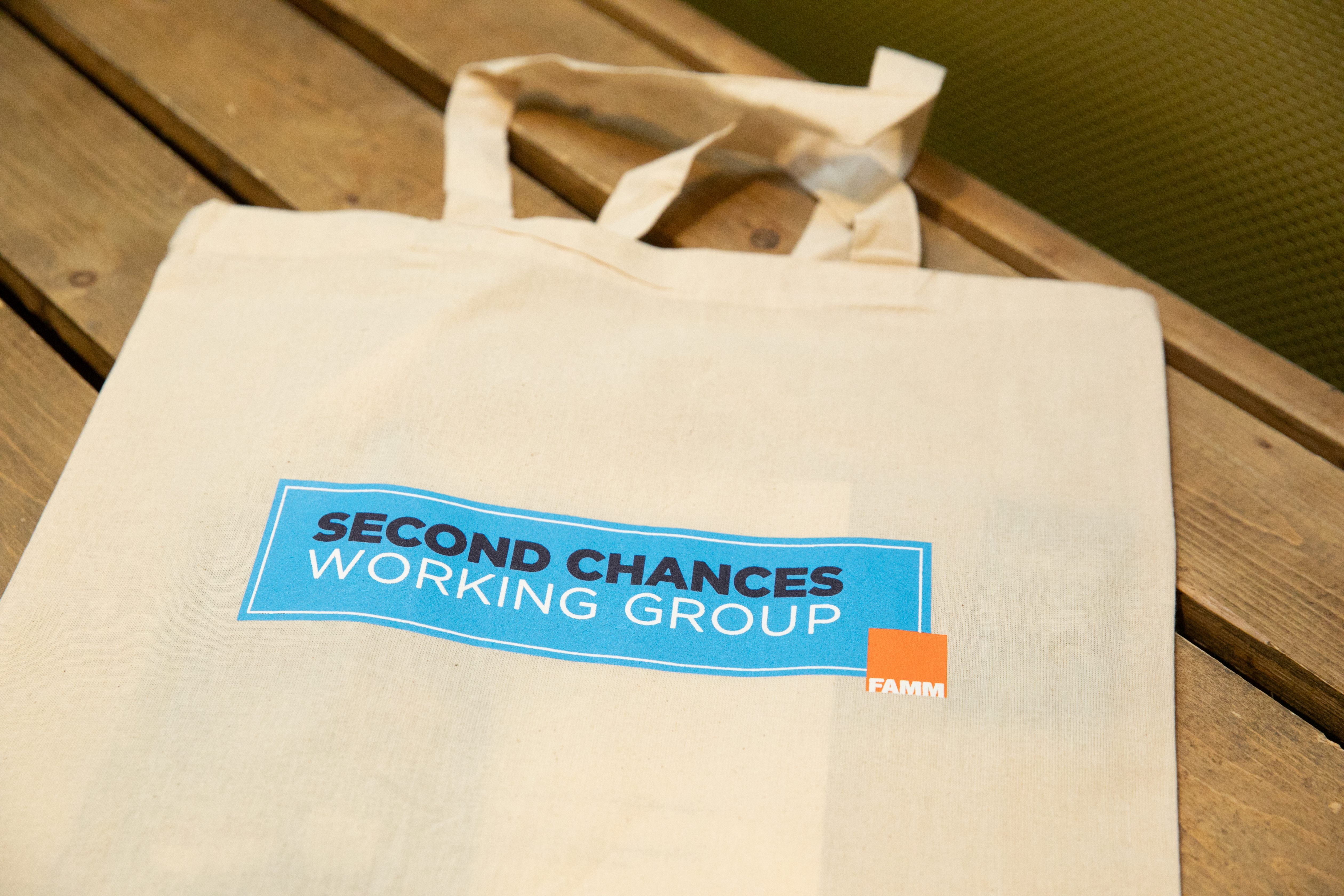 Canvas tote bag on a wooden table printed with the text “SECOND CHANCES WORKING GROUP” and a small FAMM logo.